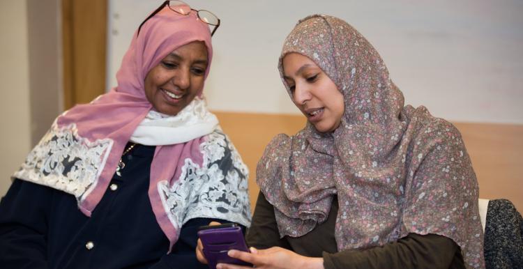 Two women sat down, both looking at a phone. They are talking to each other. Both women are wearing hijab.