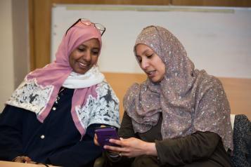 Two women sat down, both looking at a phone. They are talking to each other. Both women are wearing hijab.