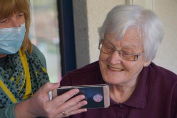 A close up of and older woman, with short white hair, smiling at a mobile phone. The phone is being held up by a younger women, wearing a surgical-style face mask.
