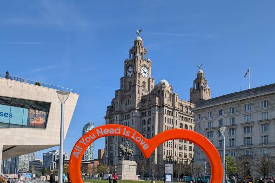 Liver building below a bright blue sky. Red heart in foreground with the words All you need is love.