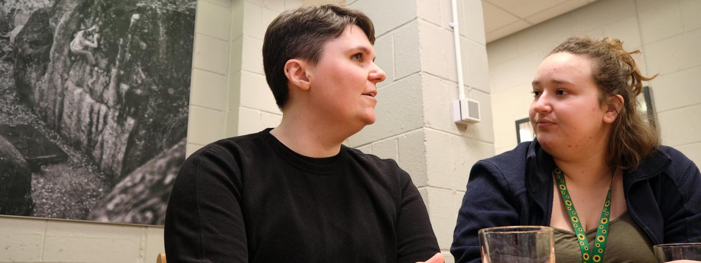 Two white people sat at a table, talking. The person on the right is wearing a sunflowers lanyard.