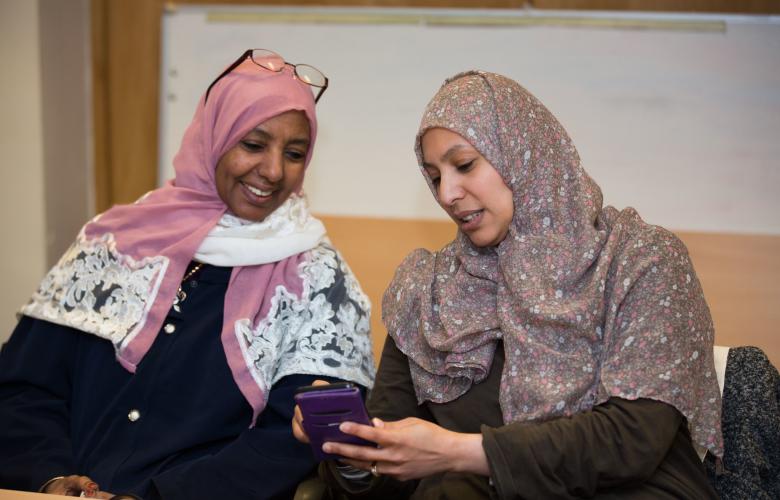 Two women sat down, both looking at a phone. They are talking to each other. Both women are wearing hijab.