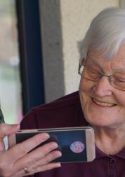 A close up of and older woman, with short white hair, smiling at a mobile phone. The phone is being held up by a younger women, wearing a surgical-style face mask.