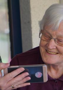 A close up of and older woman, with short white hair, smiling at a mobile phone. The phone is being held up by a younger women, wearing a surgical-style face mask.